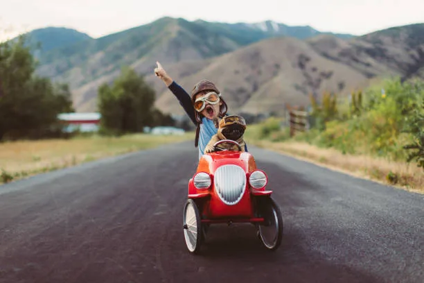 A young boy with flying goggles and flight cap races a red toy car with his pet and best friend French Bulldog along a small road in Utah, USA. Som